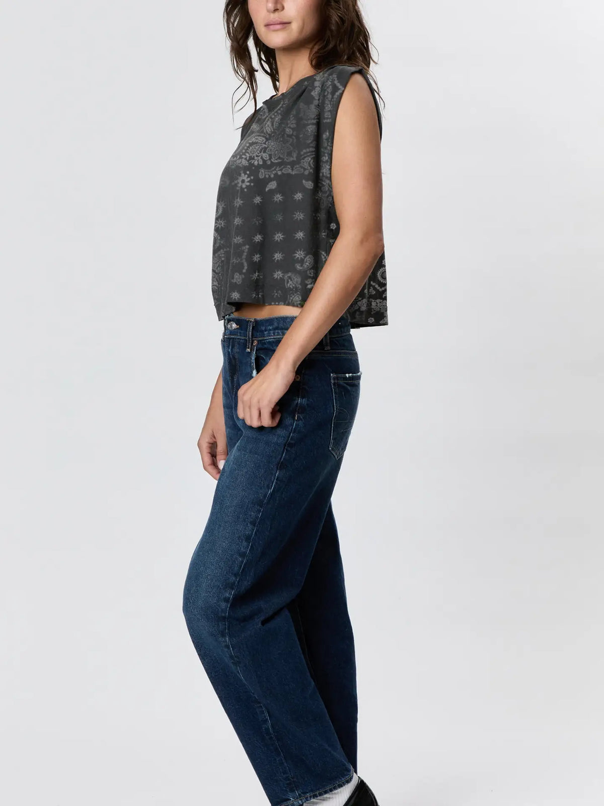 A woman stands sideways against a light background, wearing Faire's Hemp Jackie Pleat Shell Bandana—a sleeveless hemp jersey top in a dark gray pattern—paired with high-waisted blue jeans.