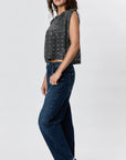 A woman stands sideways against a light background, wearing Faire's Hemp Jackie Pleat Shell Bandana—a sleeveless hemp jersey top in a dark gray pattern—paired with high-waisted blue jeans.