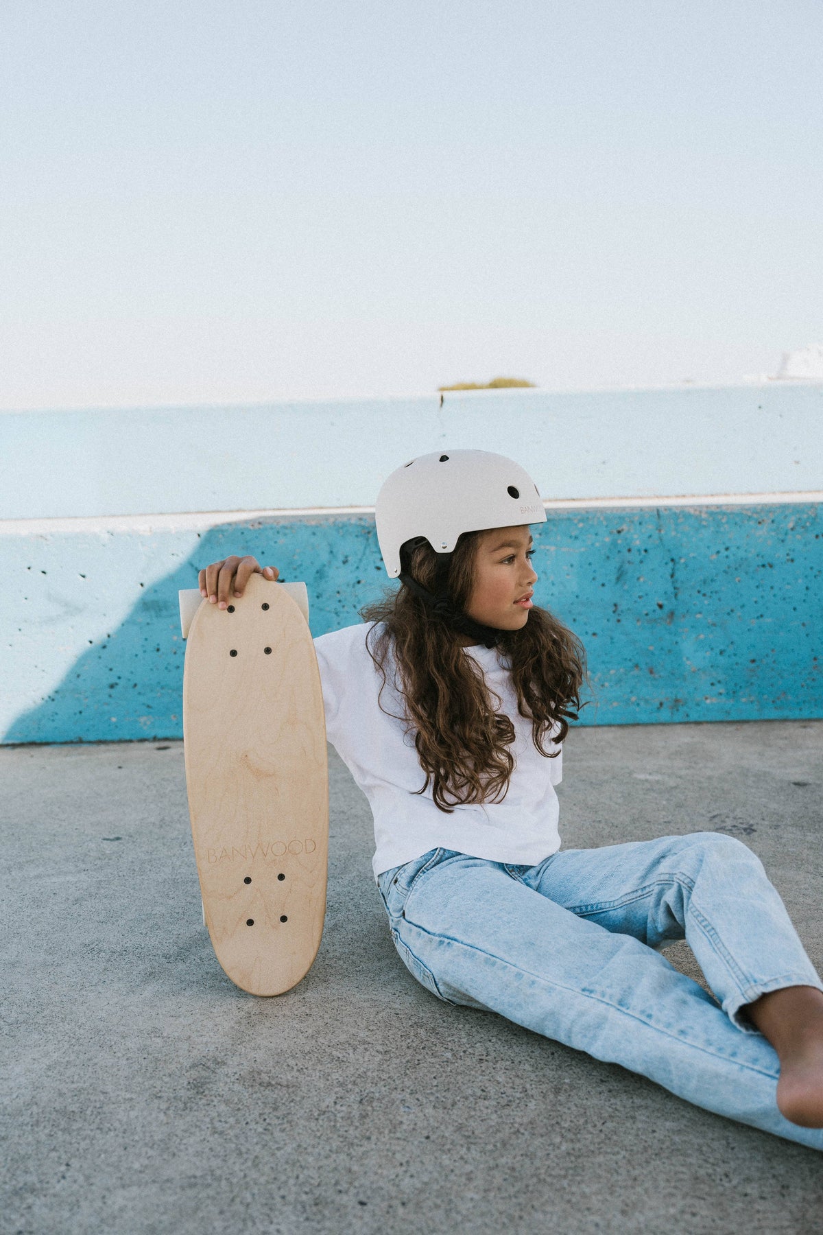 A young girl in a white helmet, t-shirt, and blue jeans sits on the ground against a blue concrete wall, holding the BANWOOD SKATEBOARD - NATURE by Faire with a Canadian maple deck under the clear sky.