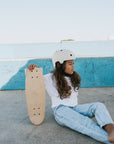A young girl in a white helmet, t-shirt, and blue jeans sits on the ground against a blue concrete wall, holding the BANWOOD SKATEBOARD - NATURE by Faire with a Canadian maple deck under the clear sky.