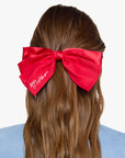 Red satin hair bow with white embroidered text from Bungalow Furniture & Accessories worn in brown hair.