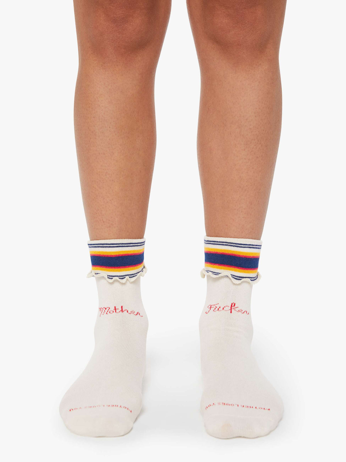 A person wearing "The Fold Please" cream-colored, ankle-length tube socks by Mother, with "Mother" and "Fucker" in pink embroidery, frilled hems, and blue, yellow, and orange striped cuffs stands on a white surface.