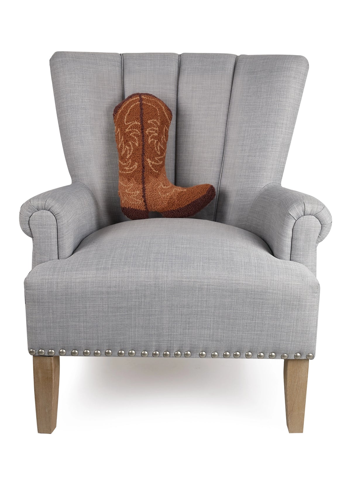 A light gray upholstered armchair with wooden legs and silver studs features the Faire Brown Cowboy Boot Pillow, a poly velvet throw pillow shaped like a brown cowboy boot, placed on the seat.