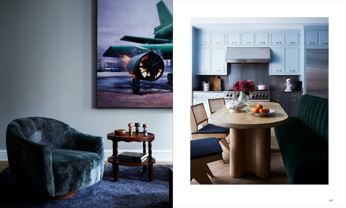 A modern space featured in "Robert Stilin New Work" by Hachette Book Group includes a dark plush chair, round table, aviation-themed art, and a kitchen with blue cabinets, stainless appliances, and a fruit- and flower-topped wood dining table.