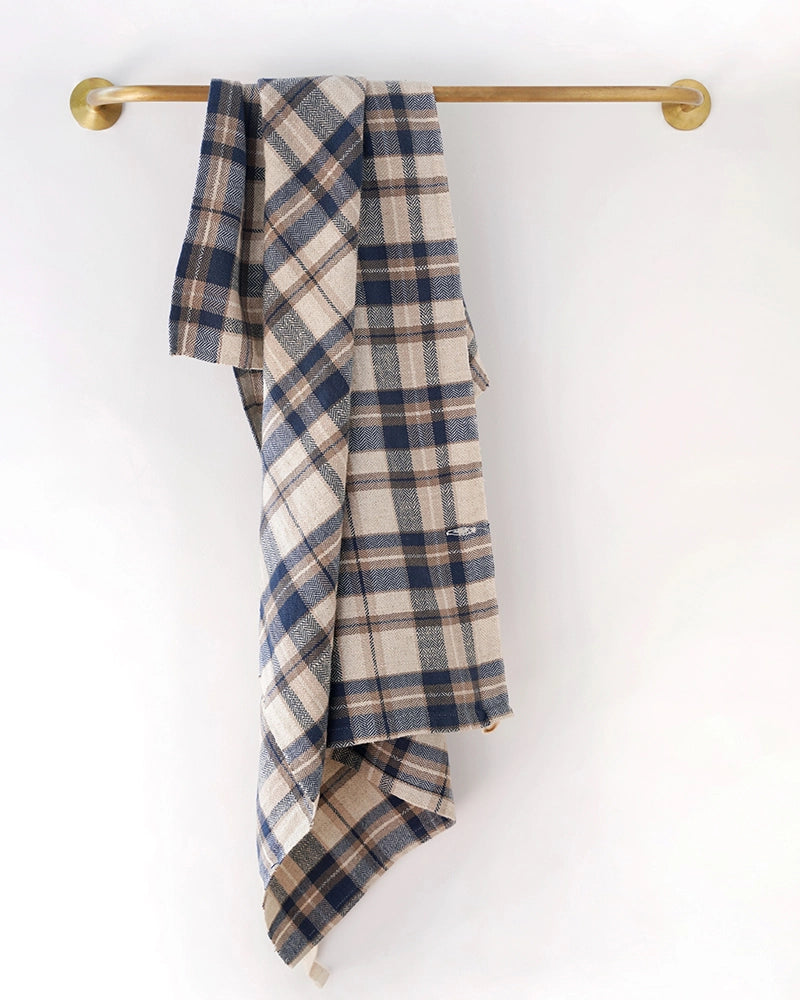 The Faire Cook's Cloth, a blue, beige, and white plaid cotton-linen blend kitchen towel, hangs on a brass towel bar against a plain white wall.