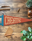 The Faire "Create" Felt Pennant rests on a wooden table with a pencil, brown envelope, cinnamon stick, crystals, and plants—ideal as creative room decor or a thoughtful gift to inspire creativity.