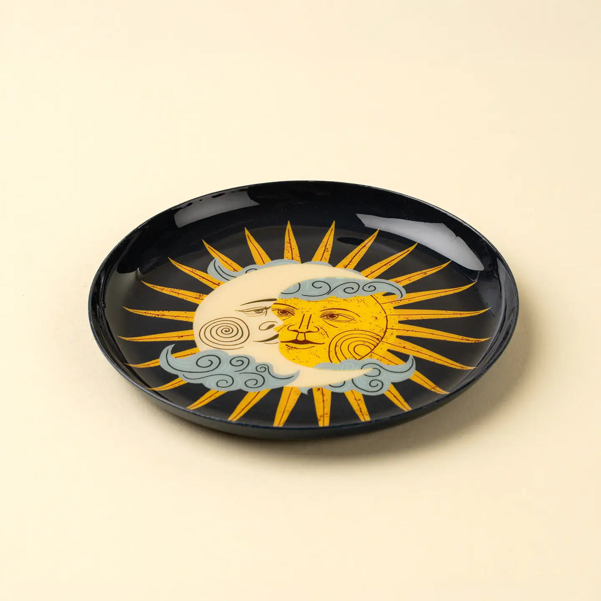 The Indaba Sun and Moon Tray is a round black accent piece featuring an artistic yellow sun and pale blue crescent moon with faces, swirling clouds, and rays on a light beige background—perfect for decorative display.