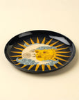 The Indaba Sun and Moon Tray is a round black accent piece featuring an artistic yellow sun and pale blue crescent moon with faces, swirling clouds, and rays on a light beige background—perfect for decorative display.
