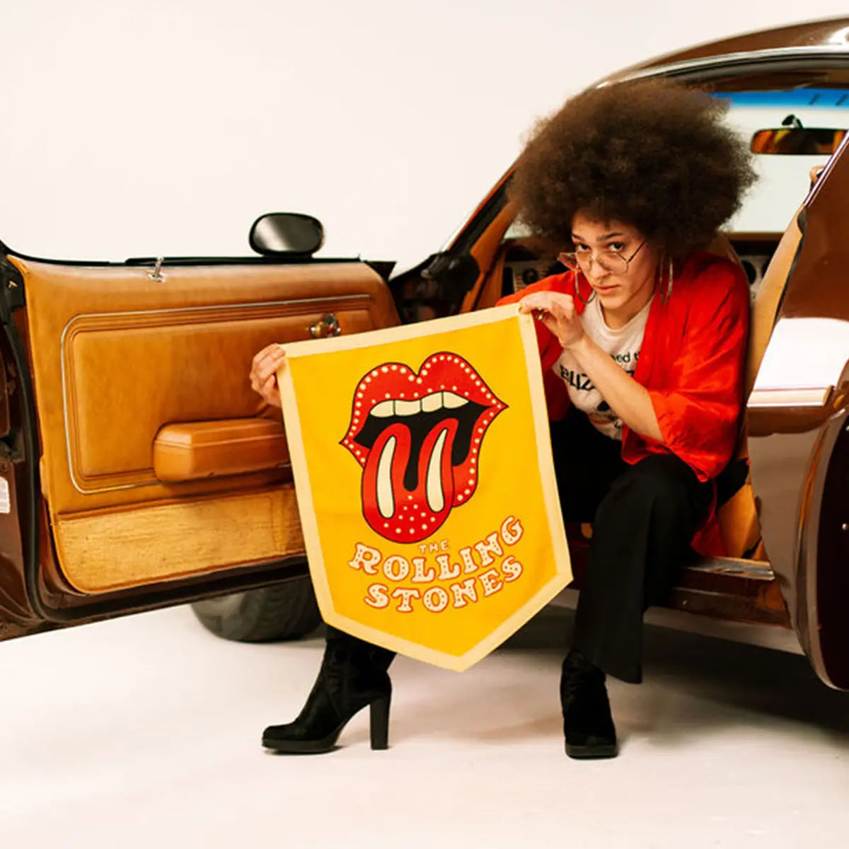 The Rolling Stones Camp Flag by Faire held by a person in black boots beside an open car door, casual outdoor style.