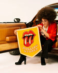 The Rolling Stones Camp Flag by Faire held by a person in black boots beside an open car door, casual outdoor style.