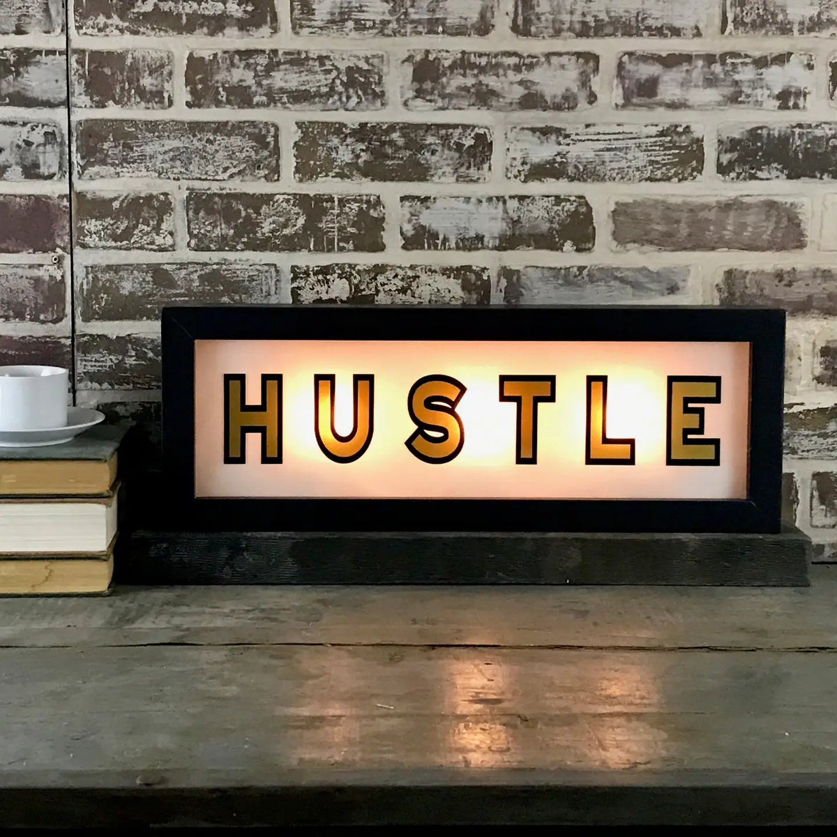 The Faire Lighted Sign featuring "HUSTLE" in bold black letters rests on a wooden surface by a brick wall, alongside stacked books and a white cup with saucer.