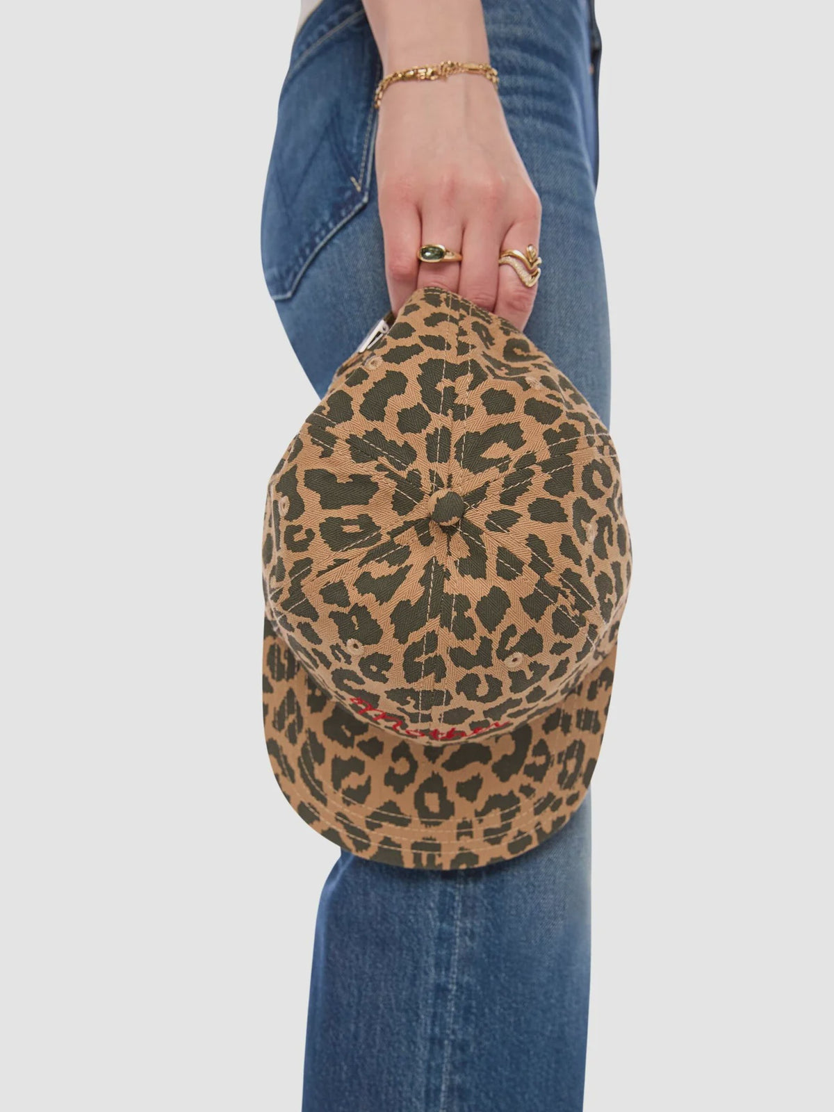 A person wearing blue jeans holds Mother’s "The Hats Off" leopard print hat by the brim, showing only their hand with wrist jewelry and part of their legs against a plain background.