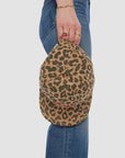 A person wearing blue jeans holds Mother’s "The Hats Off" leopard print hat by the brim, showing only their hand with wrist jewelry and part of their legs against a plain background.