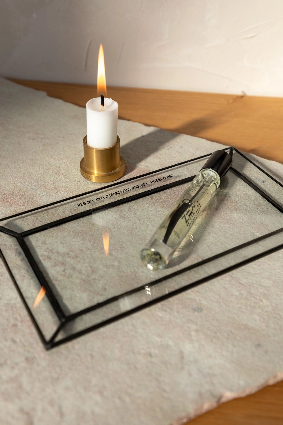 A lit white candle in a brass holder sits on a textured surface next to the Faire Glass Valet Tray, which holds a clear perfume bottle with a black cap. Warm light casts soft shadows across this elegant arrangement.