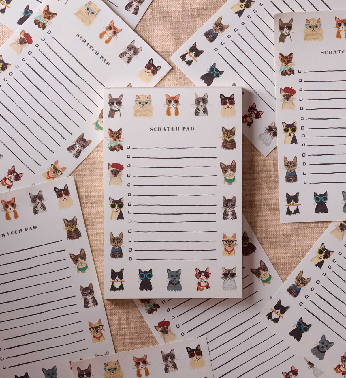The Cool Cats Notepad by Faire features illustrated tear-off sheets with colorful cats in different poses and accessories, all arranged on a textured beige surface.