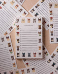 The Cool Cats Notepad by Faire features illustrated tear-off sheets with colorful cats in different poses and accessories, all arranged on a textured beige surface.