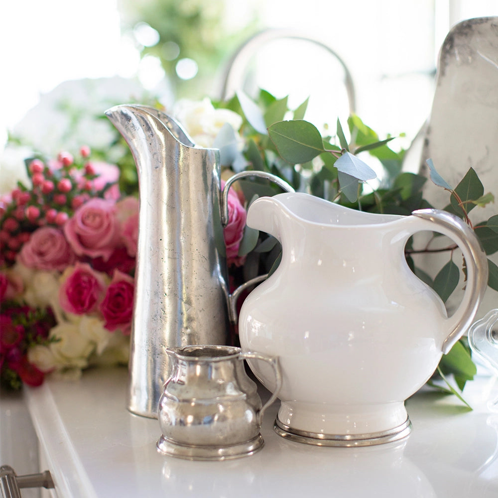 The Vintage 1815 Pitcher by Faire is displayed on a table with pink and white roses and green foliage. Soft natural light highlights the beauty and craftsmanship of this elegant piece.