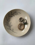 The Faire Ring Bowl, a cream hand-etched tray with a black leafy design, holds a pearl brooch and rings, serving as a chic jewelry catch-all against a white background.