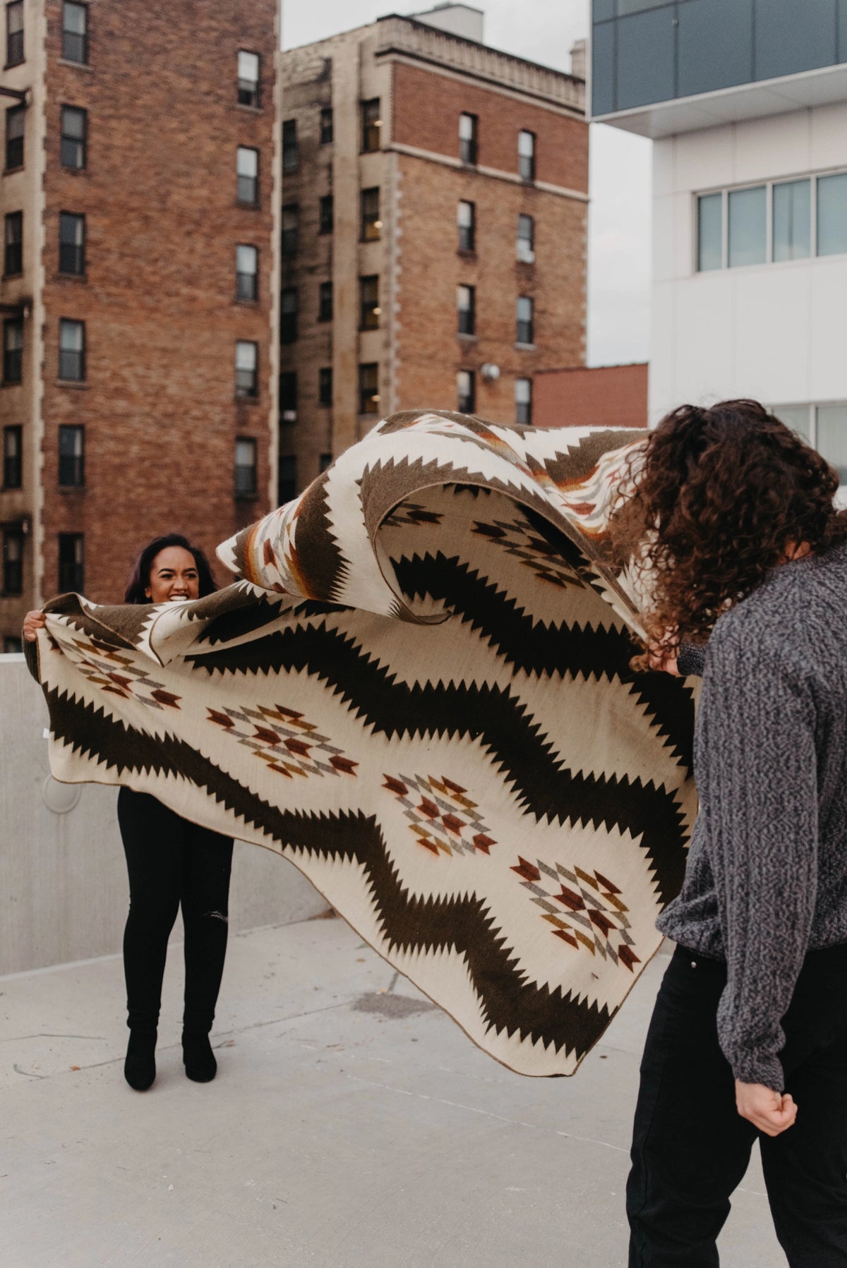 Tsachila Blanket in Chocolate, a large geometric boho woven throw held outdoors by two people.