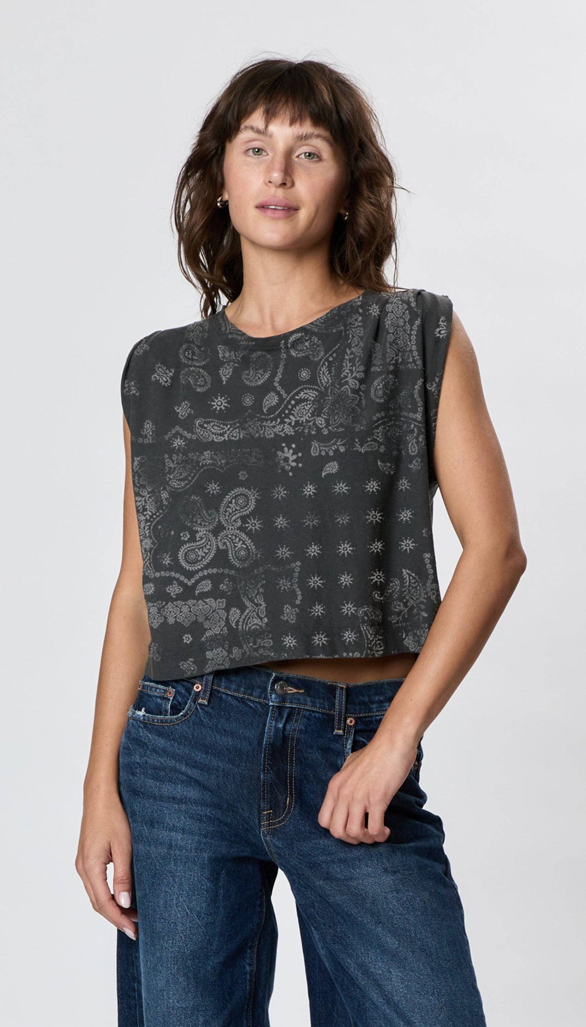 A woman with wavy brown hair wears the Faire Hemp Jackie Pleat Shell Bandana top and blue jeans, standing against a plain light gray background. She looks at the camera with a relaxed expression and one hand by her side.