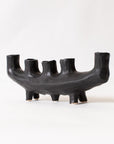 The Faire Black Crescent Shape is a sculptural, matte black ceramic candle holder with five uneven cylindrical slots and four short legs, shown against a plain white background.