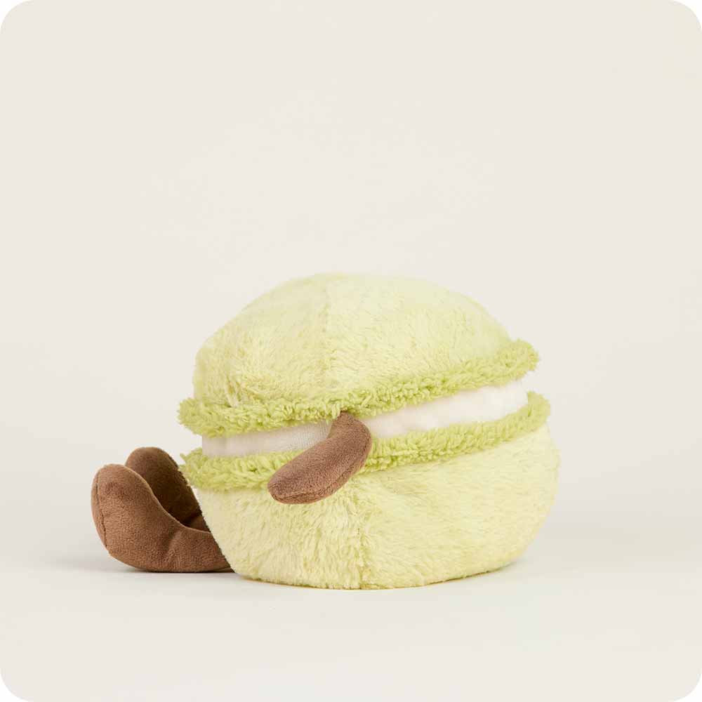 The Faire Green Macaroon Warmie is a plush toy shaped like a light green macaron with brown, floppy arms and legs, white filling, and is microwavable for sensory comfort, shown on a plain light background.