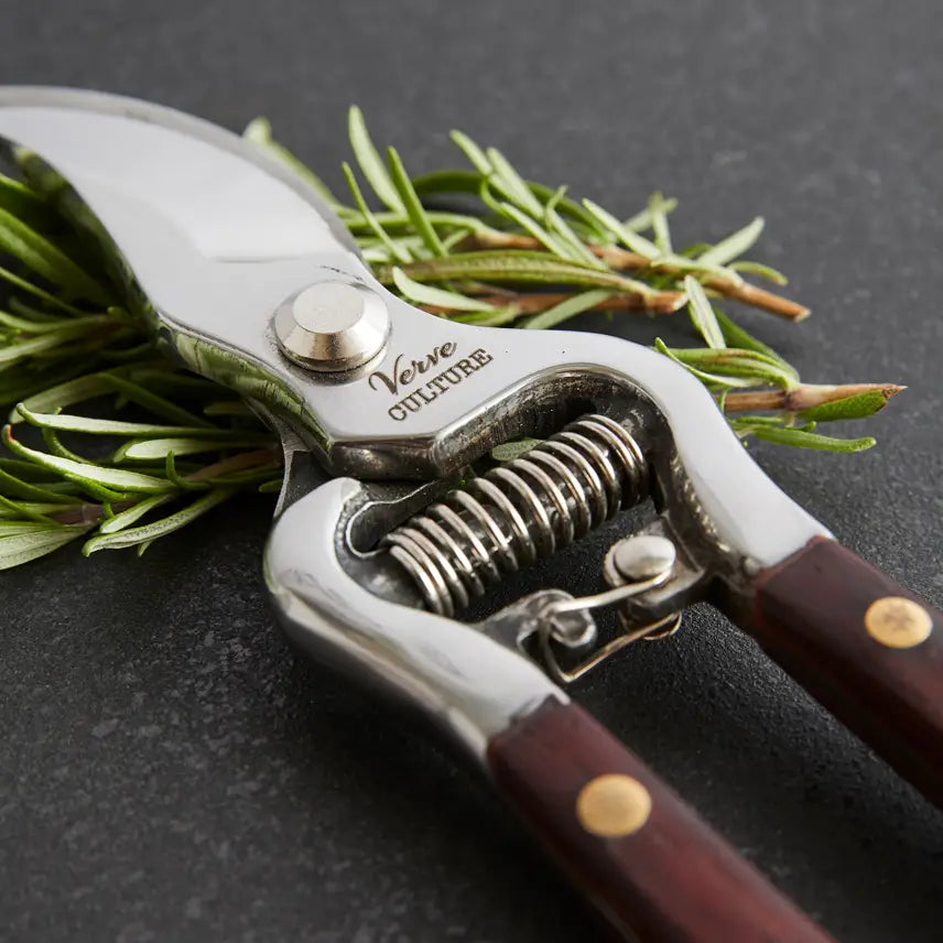 The Faire Thai Kitchen Garden Shears, featuring wooden handles and stainless steel blades, rest on a dark surface as they trim fresh rosemary.