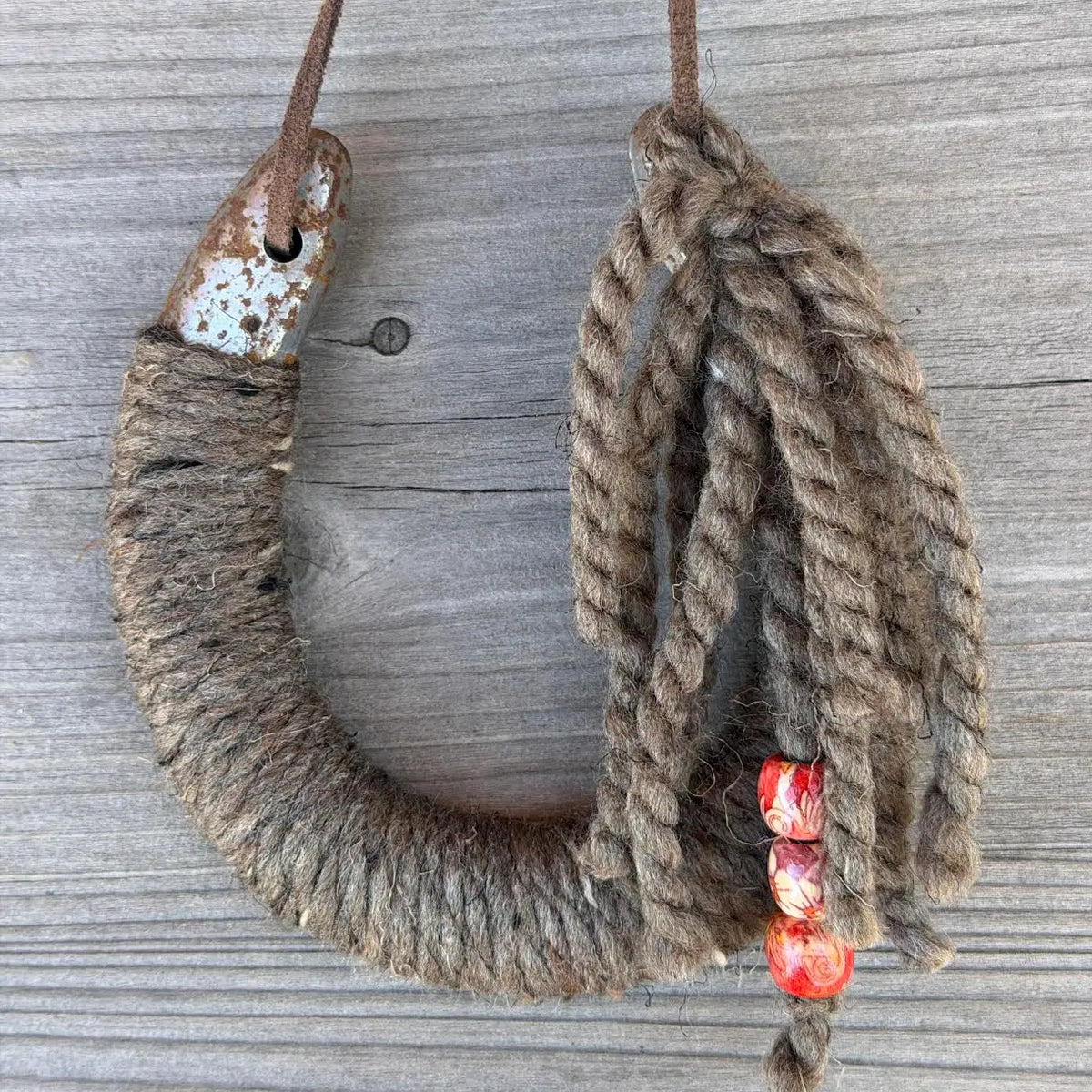 The Lucky Horseshoe by Faire is a large, upcycled horseshoe-shaped hook wrapped in coarse rope against weathered wood. Loose rope strands end with three red and orange patterned beads—ideal for rustic decor.