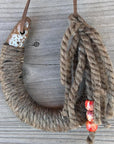 The Lucky Horseshoe by Faire is a large, upcycled horseshoe-shaped hook wrapped in coarse rope against weathered wood. Loose rope strands end with three red and orange patterned beads—ideal for rustic decor.