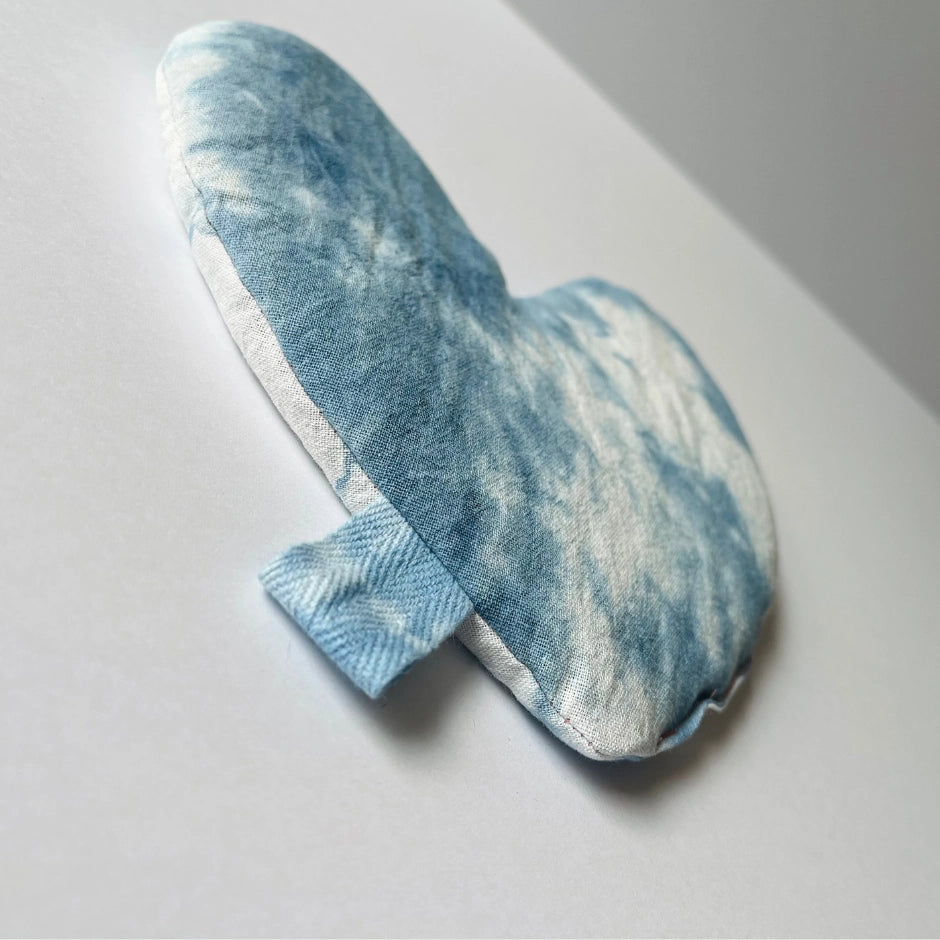 The Faire Heart Eye Pillow, featuring a heart shape and blue-and-white tie-dye cotton, rests on a white surface. A small blue fabric tab extends from the side—ideal for gentle anxiety relief.