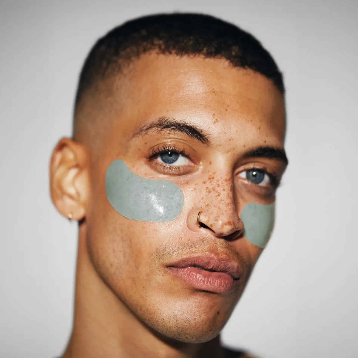 A person with short hair, blue eyes, and freckles wears Faire's Hyper Eyes Mask green under-eye patches to depuff and brighten eyes. They have small hoop earrings and a nose ring, looking at the camera against a neutral background.