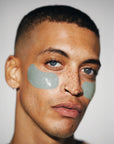 A person with short hair, blue eyes, and freckles wears Faire's Hyper Eyes Mask green under-eye patches to depuff and brighten eyes. They have small hoop earrings and a nose ring, looking at the camera against a neutral background.