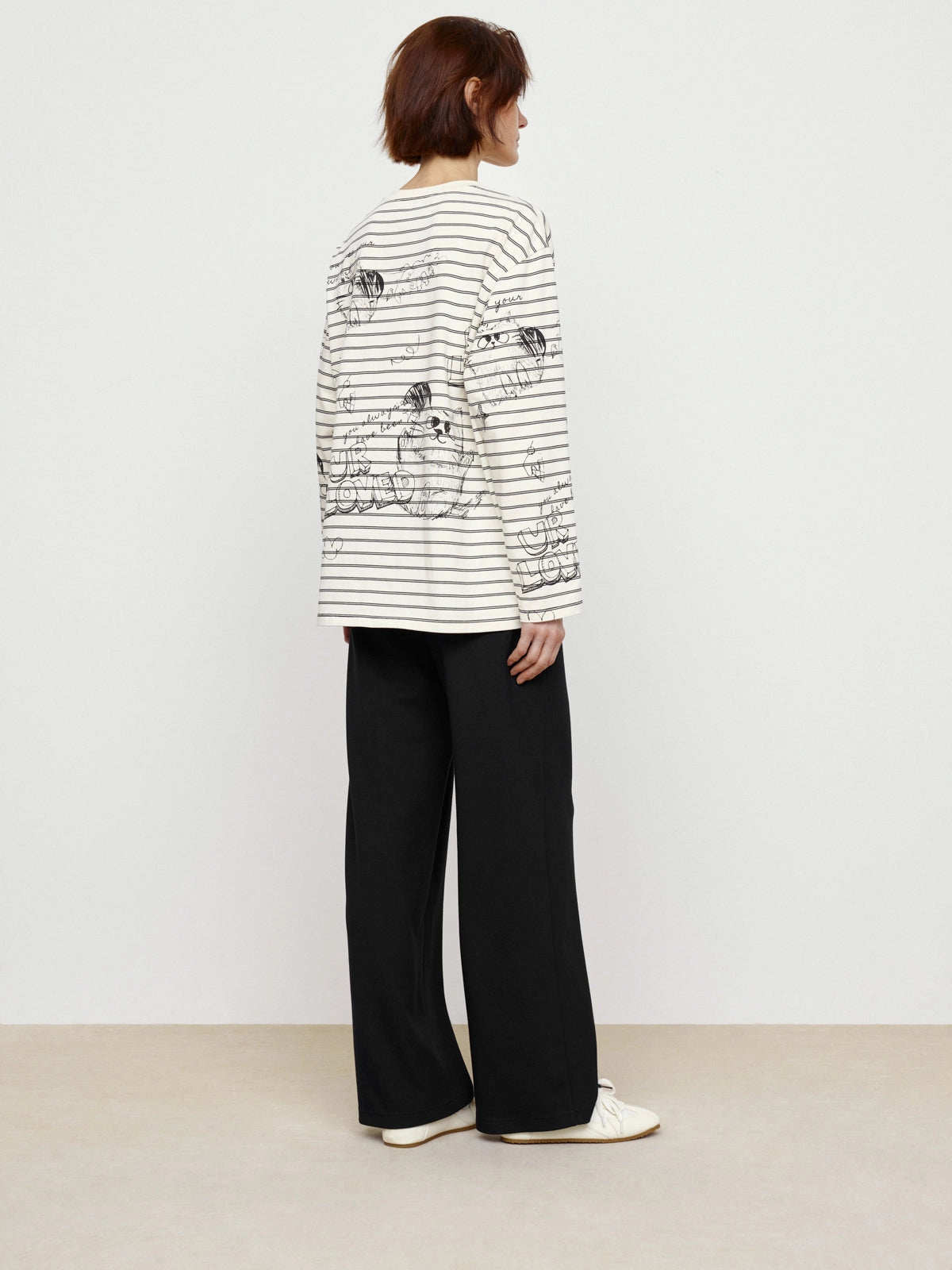 A person with short brown hair stands sideways, wearing Faire’s Graffiti Stripe Tee with black stripes and sketch-style flowers, paired with loose black pants and white shoes.