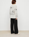 A person with short brown hair stands sideways, wearing Faire’s Graffiti Stripe Tee with black stripes and sketch-style flowers, paired with loose black pants and white shoes.