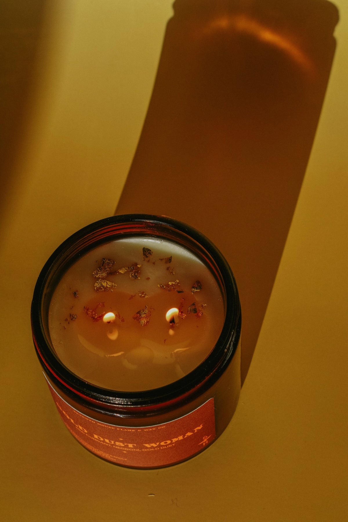 Brown jar Gold Dust Woman Candle by Faire with flower petals on a yellow surface, casting a long shadow.