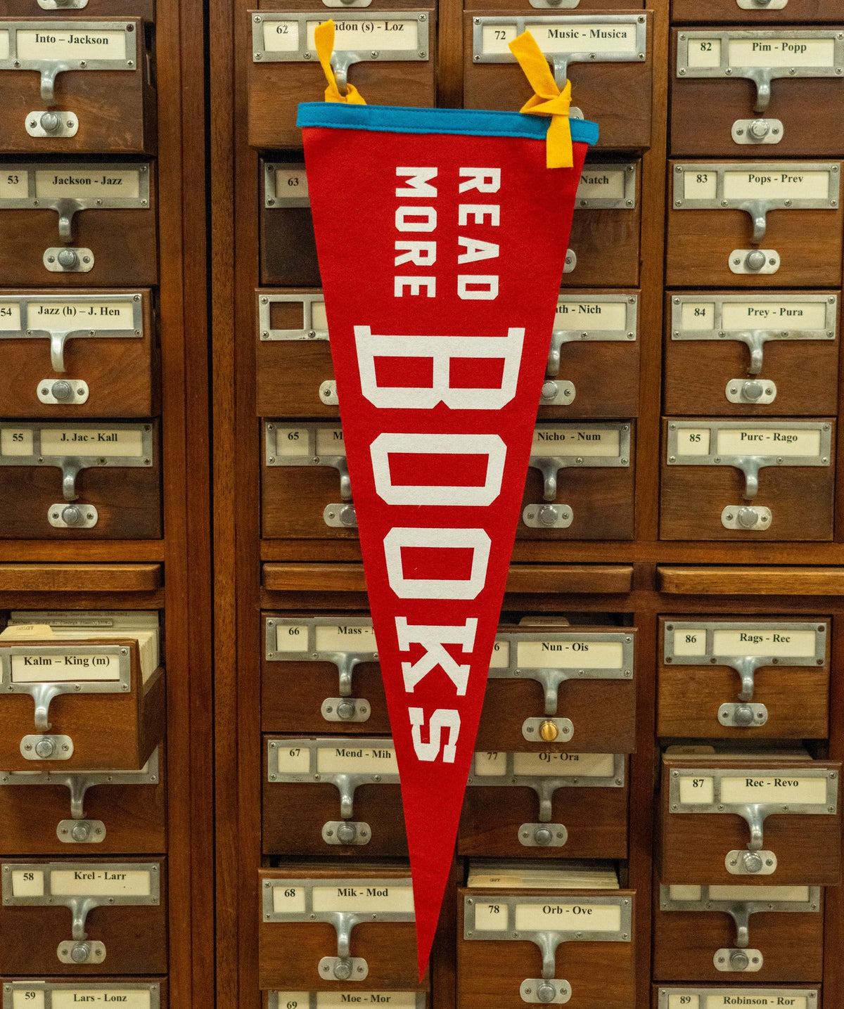 The Faire Read More Books Pennant features red felt with "READ MORE BOOKS" in white letters and blue and yellow ribbons, hanging on a wooden card catalog to inspire book lovers.