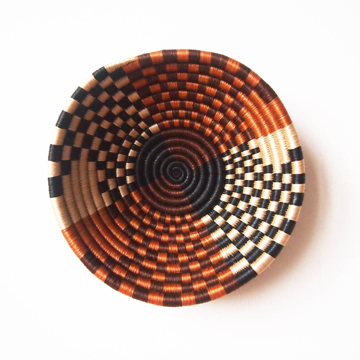 The Muko Small Bowl by Faire is a handmade Rwandan basket with a geometric spiral in black, brown, cream, and orange tones, displayed on a white background.