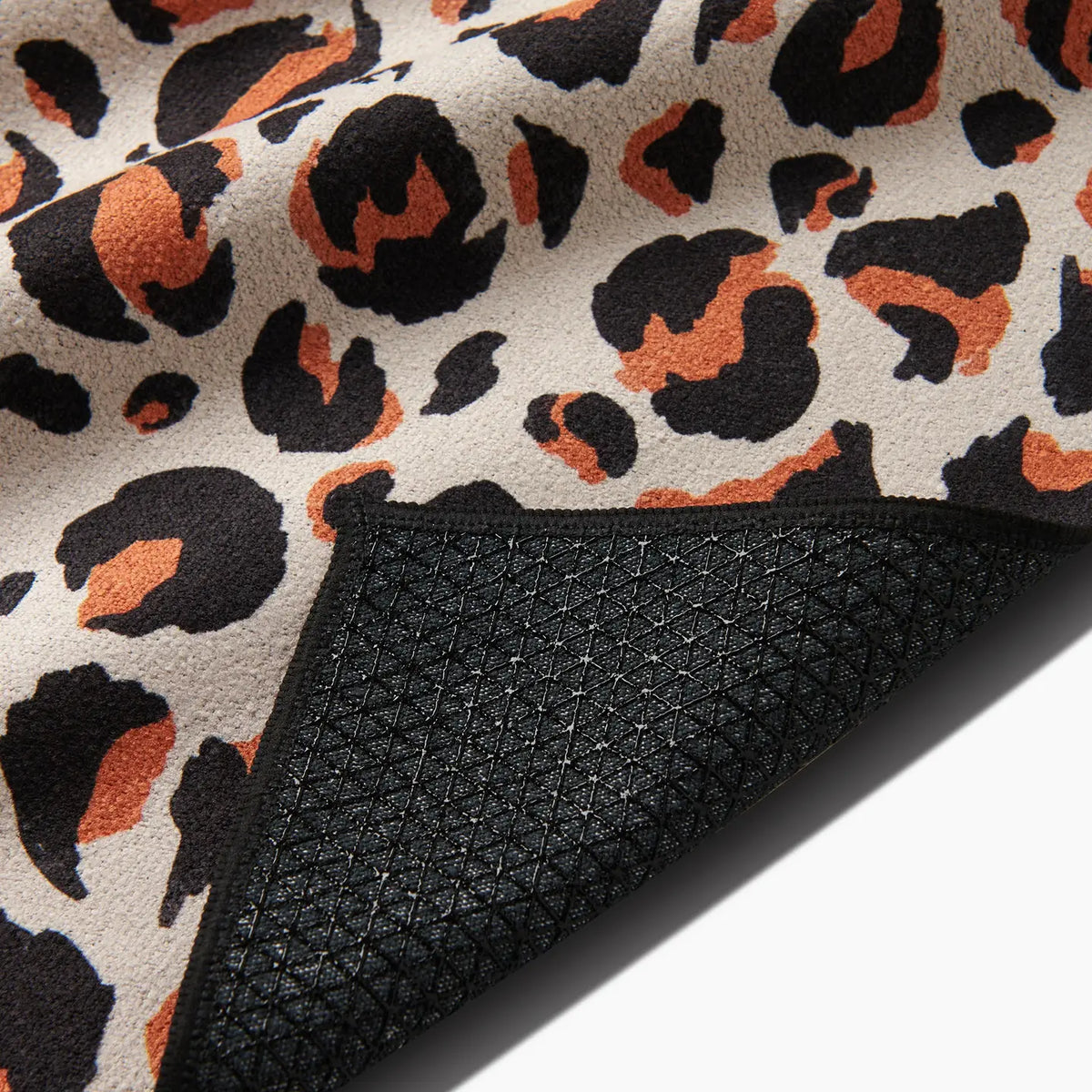 A close-up of the Faire Leopard Yoga Towel shows its brown, black, and orange leopard print. The flipped corner reveals a textured black, slip-resistant backing for secure grip.