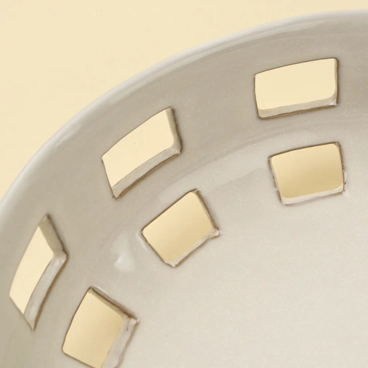 Close-up of the Indaba Cypress Fruit Bowl L, a white ceramic kitchen piece featuring square cut-out patterns along the rim, displayed on a light beige countertop.