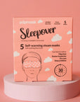 A pink box of "Faire eye masks" contains 5 rose-scented self-warming steam masks for migraines, tension headaches, and dry eyes relief. Positioned on a circular platform with a pink background, they're perfect for your next jet setter adventure!.