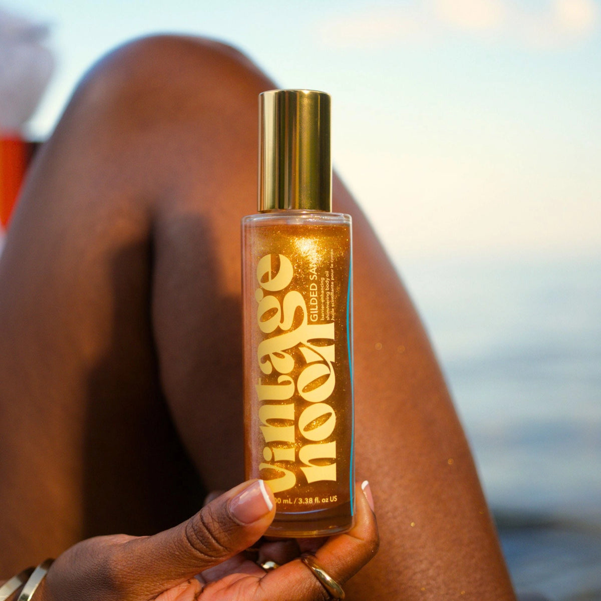 A hand holds a bottle of Faire Shimmer Body Oil—a nourishing, antioxidant-rich golden formula—against a backdrop featuring a person’s leg and an out-of-focus outdoor setting, possibly near water.
