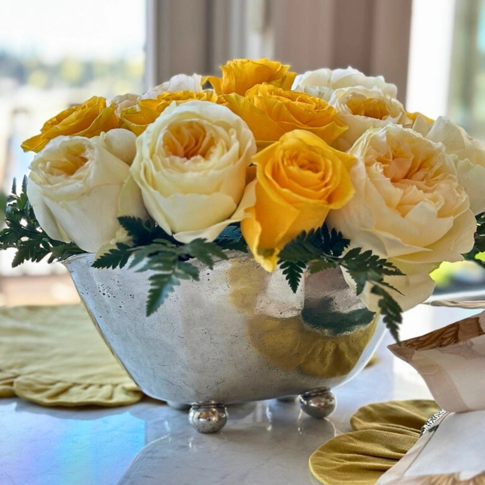 A Vintage Scalloped Bowl by Faire, filled with yellow and white roses and green fern leaves, sits on a table with yellow placemats beside a sunlit window.