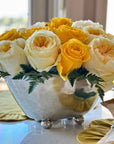 A Vintage Scalloped Bowl by Faire, filled with yellow and white roses and green fern leaves, sits on a table with yellow placemats beside a sunlit window.