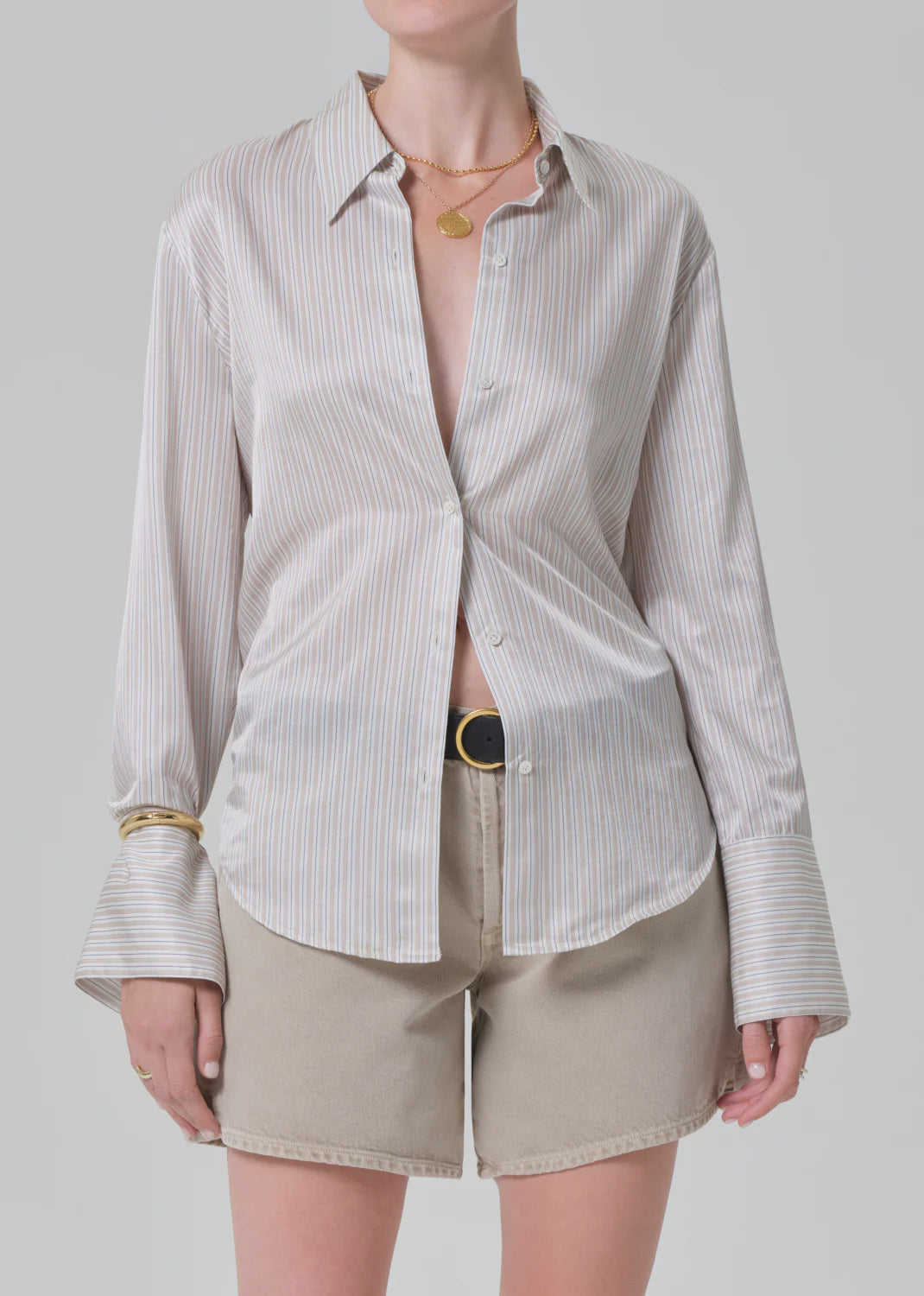 Wearing the camilia shirt S26 by Citizens Of Humanity/AGOLDE—a shiny beige striped cotton-silk button-down with flared cuffs—paired with beige shorts, a black belt, gold necklaces, and a bracelet against a light gray background.