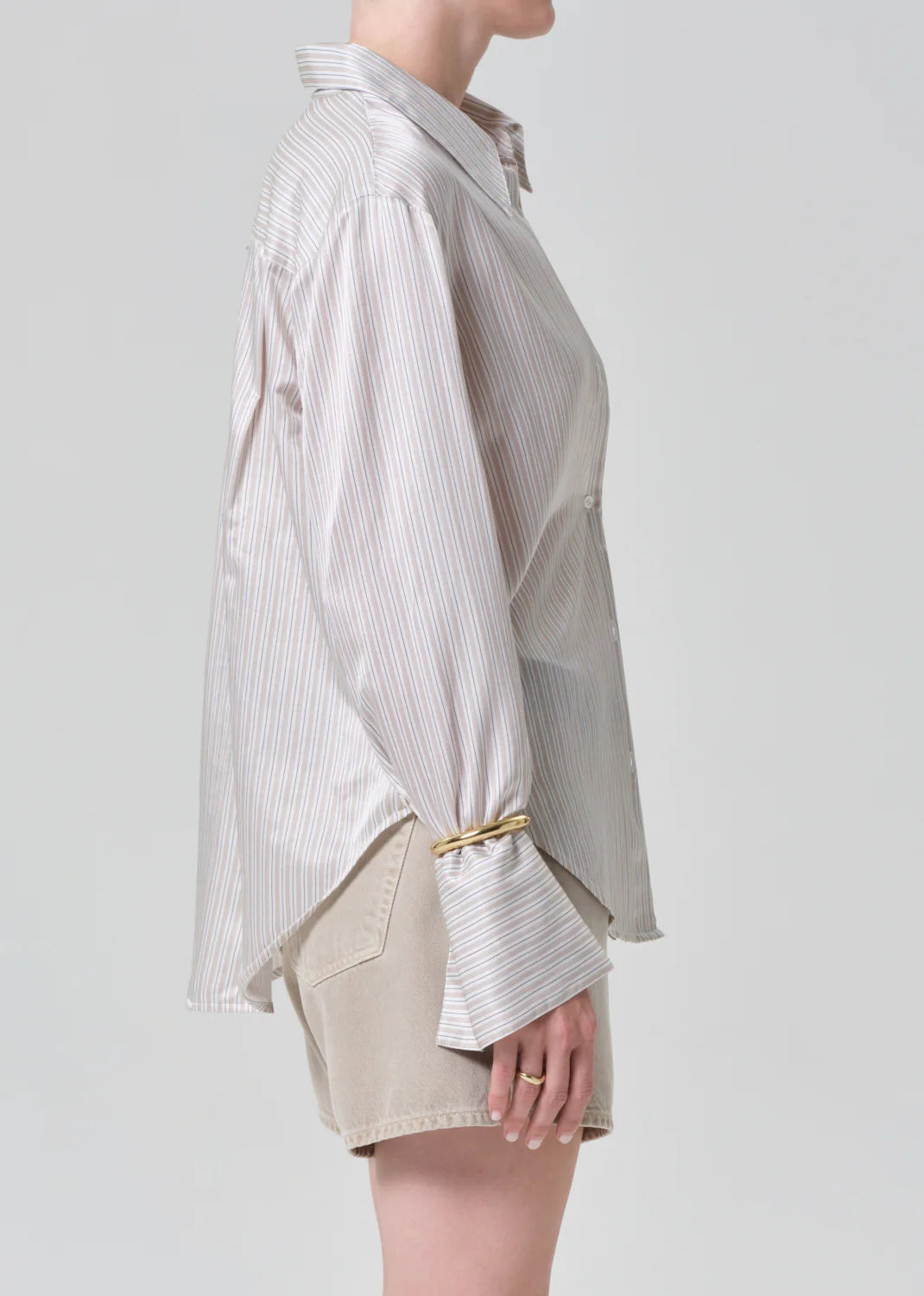 A person in profile wears the camilia shirt S26 by Citizens Of Humanity/AGOLDE—a loose, long-sleeve cotton silk blend top with flared cuffs—paired with beige shorts, standing against a plain light background.