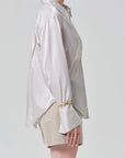 A person in profile wears the camilia shirt S26 by Citizens Of Humanity/AGOLDE—a loose, long-sleeve cotton silk blend top with flared cuffs—paired with beige shorts, standing against a plain light background.