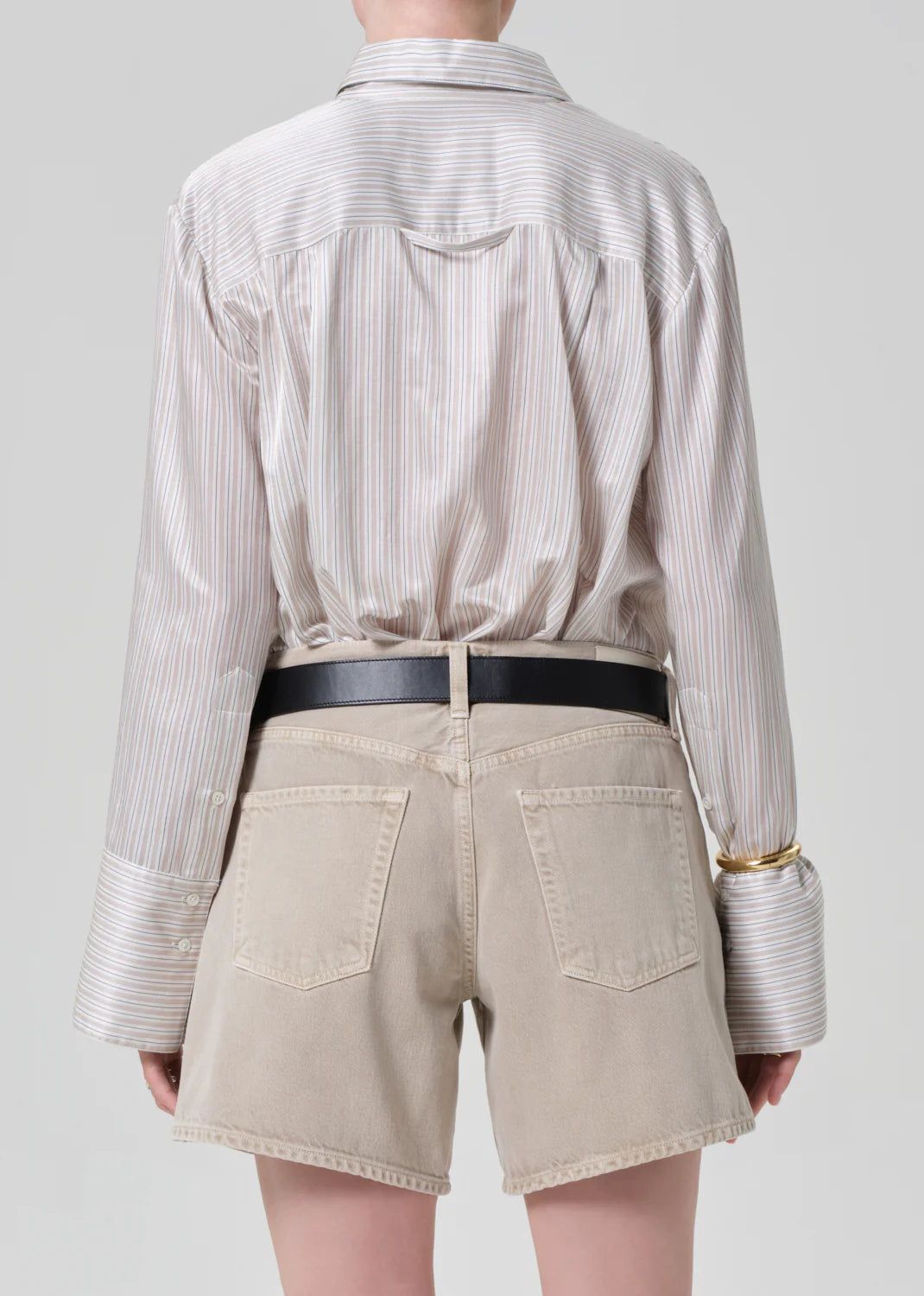 A person is seen from behind wearing the Citizens Of Humanity/AGOLDE camilia shirt S26, a beige striped cotton silk blend with oversized cuffs, tucked into light beige denim shorts and styled with a black belt and gold bangle.