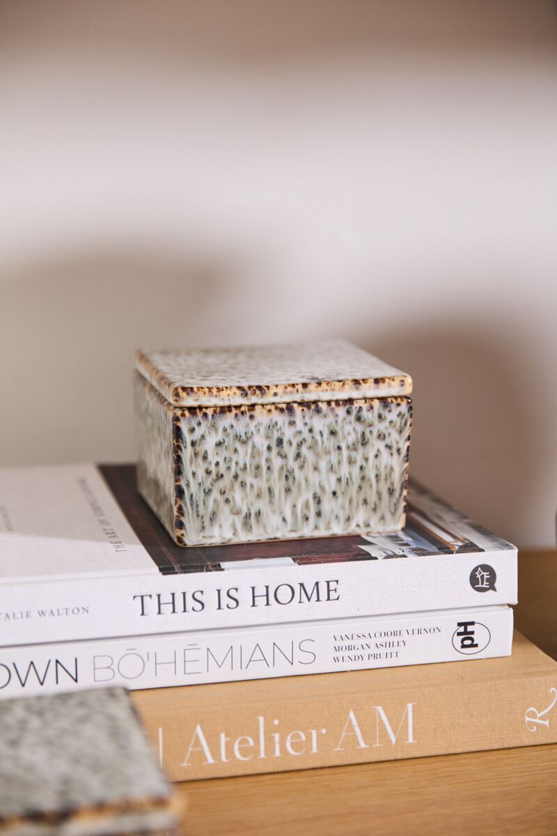 The Whisper Lidded Box by Accent Decor, a square textured ceramic piece, rests atop "THIS IS HOME" and "BOHÉMIANS," providing stylish jewelry storage and serving as a chic decorative accent on a wooden surface with a neutral backdrop.