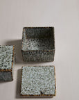 The Whisper Lidded Box by Accent Decor, a square ceramic piece with a speckled textured glaze, comes with two matching lids. Open to display its interior, it’s perfect for storing jewelry; one lid is shown resting beside it on a light surface.