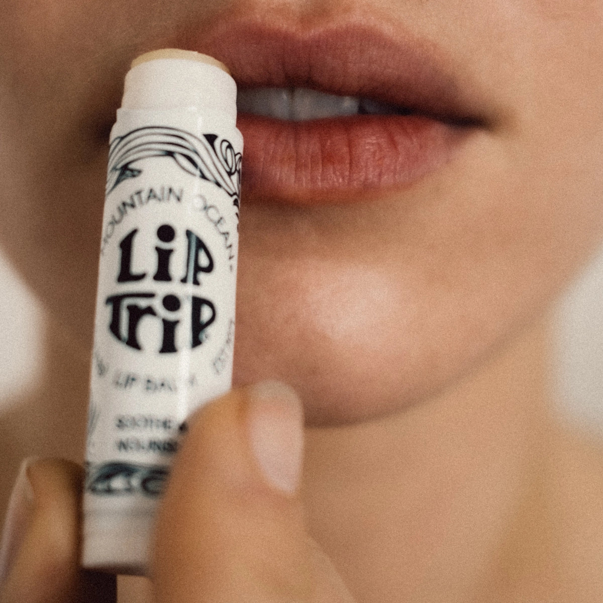 A close-up of a person holding an uncapped tube of Faire Skin Trip Lip Balm near their parted lips, ready to apply the moisturizing balm.
